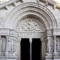 Ce portail sculpté est ajouté à l’église entre 1180 et 1190. Avec la façade de l’abbaye de Saint-Gilles qui lui est très légèrement antérieure, il constitue un des deux plus grands ensembles sculptés de l’art roman en Provence. Le tympan de Saint-Trophime reprend le thème biblique du tétramorphe évoquant la vision d'Ézéchiel ou l'Apocalypse de saint Jean, symbole ensuite des quatre Évangélistes ; il montre un Christ triomphant et justicier, assis, tenant sur ses genoux la bible et bénissant avec ses deux doigts de sa main droite levée. Il est entouré par les symboles classiques des quatre évangélistes : un lion ailé pour saint Marc, un ange (ou un homme ailé) pour saint Mathieu, un aigle pour saint Jean et un taureau ailé pour saint Luc. Les deux évangélistes figurant au bas du tympan Marc et Luc, qui à la différence de Mathieu et Jean n'ont pas connu le Christ, ne regardent pas le fils de Dieu.